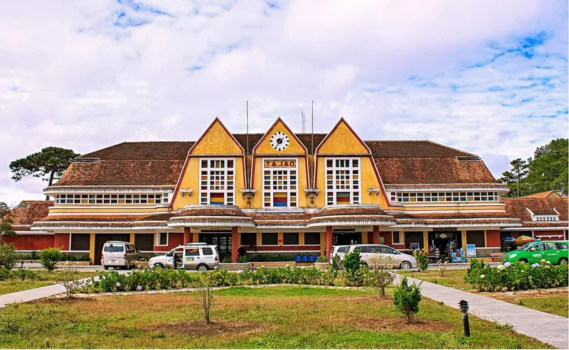 Mặt tiền vàng ba mái nhọn của nhà ga Đà Lạt nổi bật dưới bầu trời nhiều mây với khu vườn phía trước.