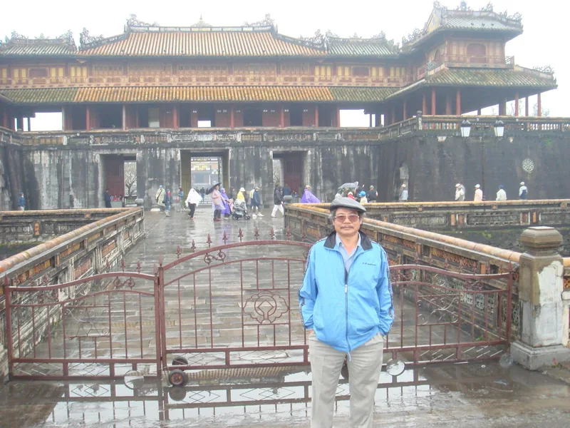 Một người đàn ông áo khoác xanh đứng trên cầu trước cổng Ngọ Môn ở Đại Nội Huế vào ngày mưa.