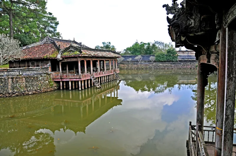 Một nhà thủy tạ bằng gỗ trên cột soi bóng xuống hồ nước tĩnh lặng trong khu lăng tẩm cổ ở Huế.