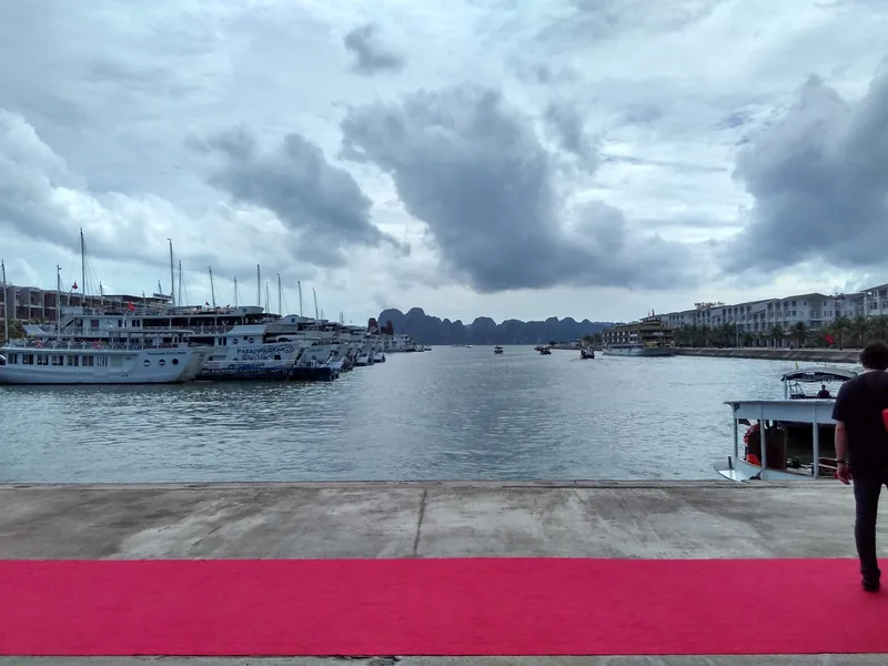 Tấm thảm đỏ dẫn ra cầu cảng với các tàu du lịch, phía xa là những núi đá vôi vịnh Hạ Long dưới bầu trời nhiều mây.